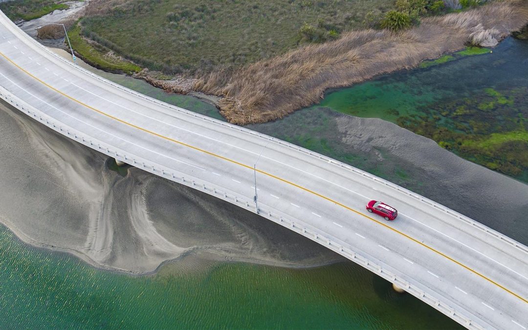 Photograph of birds eye view of SUV with surf board on roof crossing marsh bridge taken with a drone